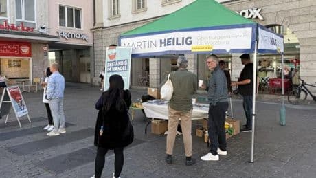 Ein Sonnenschirm am Parteistand. Die grünen Logos überdeckt mit blauen Streifen